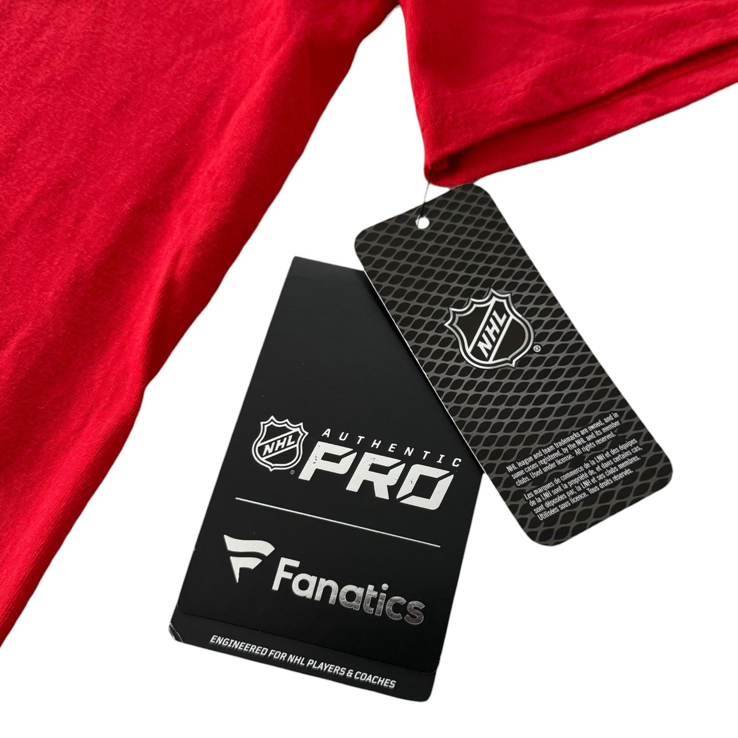 A close-up of a red garment with two hanging tags, one black NHL Authentic Pro Fanatics tag and one black NHL hangtag, on a white background.