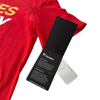 A close-up of a red short-sleeve T-shirt with partial yellow and white lettering visible, featuring a black Fanatics hangtag attached near the shoulder on a white background.