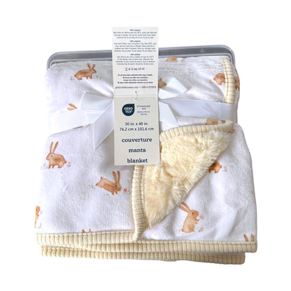 A folded baby blanket with a white sherpa fleece lining and a cream ribbed cotton cover, printed with a brown bunny pattern. The blanket is tied with a white ribbon and a paper tag from the brand Gerber