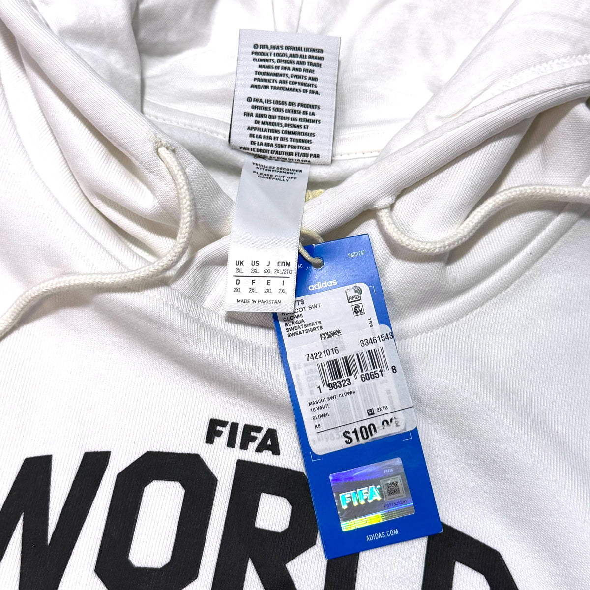 Close-up of a white Adidas hoodie with drawstrings, black printed text, and attached retail tags including a blue FIFA hangtag and a size chart tag.