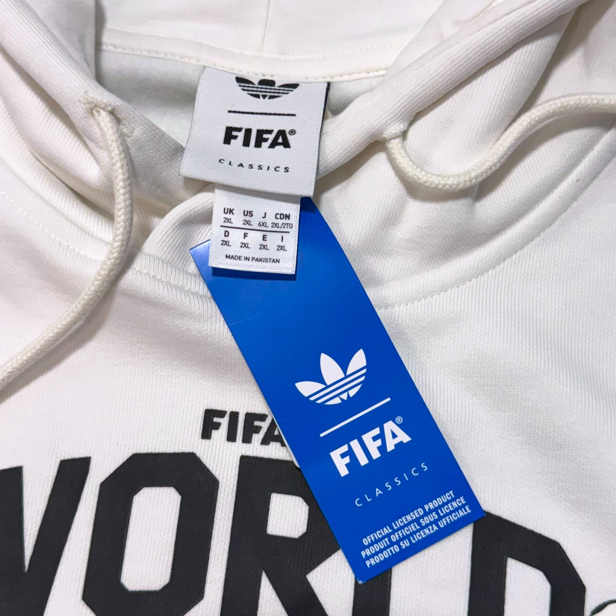 Close-up of a white Adidas x FIFA Classics hoodie showing the neck label, blue hangtag, and part of the black printed 'FIFA WORLD' text on the chest.