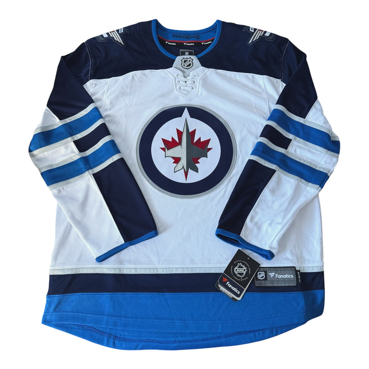 A Winnipeg Jets hockey jersey in white, navy, and blue, featuring the team's circular logo on the chest and two brand tags near the hem.